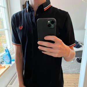 Svart pikétröja från Fred Perry - Säljer en snygg svart pikétröja från Fred Perry. Den har klassiska ränder i orange och vit på krage och ärmslut, samt Fred Perry-loggan broderad på bröstet. Perfekt för en stilren look, skit snygg!!