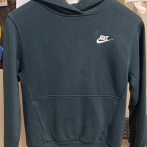 Mörkgrön hoodie från Nike - Säljer en snygg mörkgrön hoodie från Nike i storlek M.(137-147). Den är perfekt för höst och vinter med sin mysiga och bekväma passform. Hoodien har en klassisk design med Nike-loggan i vitt på bröstet. Perfekt för både träning och vardag!