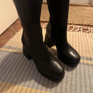 Svarta boots med klack - Snygga svarta boots i skinn med en rejäl klack och dragkedja på sidan. Perfekta för höst och vinter, dessa boots ger en stilren look och är superbekväma att gå i. De har en modern design med en fyrkantig tå som ger extra edge till din outfit.