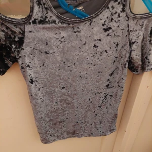 Grå sammetstopp från Flowers by Zoe - Snygg grå sammetstopp från Flowers by Zoe med cold shoulder-design. Perfekt för en kväll ute eller en fest. Toppen har en glansig finish och korta ärmar. Den är både bekväm och stilren, vilket gör den till ett måste i garderoben.