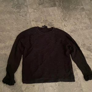 Svart stickad tröja med volangdetaljer - Säljer en svart stickad tröja med långa ärmar och snygga volangdetaljer vid ärmsluten. Perfekt för höst och vinter, den är både stilren och mysig. Passar bra till både vardag och fest. . Den är i storlek M men passar även S