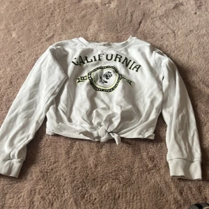 Vit långärmad tröja från H&M med tryck - Säljer en superfin vit långärmad tröja från H&M. Den har ett coolt 'California' tryck med en ros och texten 'Athletic First League' i svart och grönt. Tröjan är croppad och har en knytning framtill, vilket ger den en avslappnad och trendig look. Perfekt för vår och sommar!