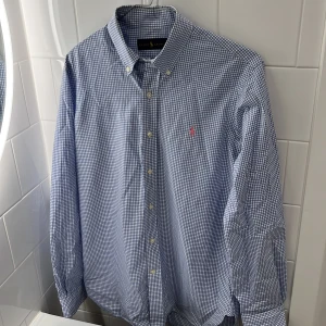 Blå och vit rutig skjorta från Ralph Lauren - Säljer en snygg blå och vit rutig skjorta från Ralph Lauren. Skjortan har långa ärmar och klassiska knappar framtill. Den är i bomull och har en broderad rosa logga på bröstet. Bra skick, 8/10. Lång modell på skjortan så passar bra om du är lång. M/L