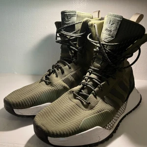 Adidas F/1.3 Primeknit Boots ‘Olive Cargo’ (EU 46,5) - Stilrena och slitstarka Adidas ATRIC F/1.3 Primeknit Boots i färgen ‘Olive Cargo’, storlek EU 46,5. Dessa boots kombinerar urban streetwear med funktionella outdoor-detaljer, perfekta för både stad och friluftsliv. Primeknit-ovandelen ger en bekväm, 