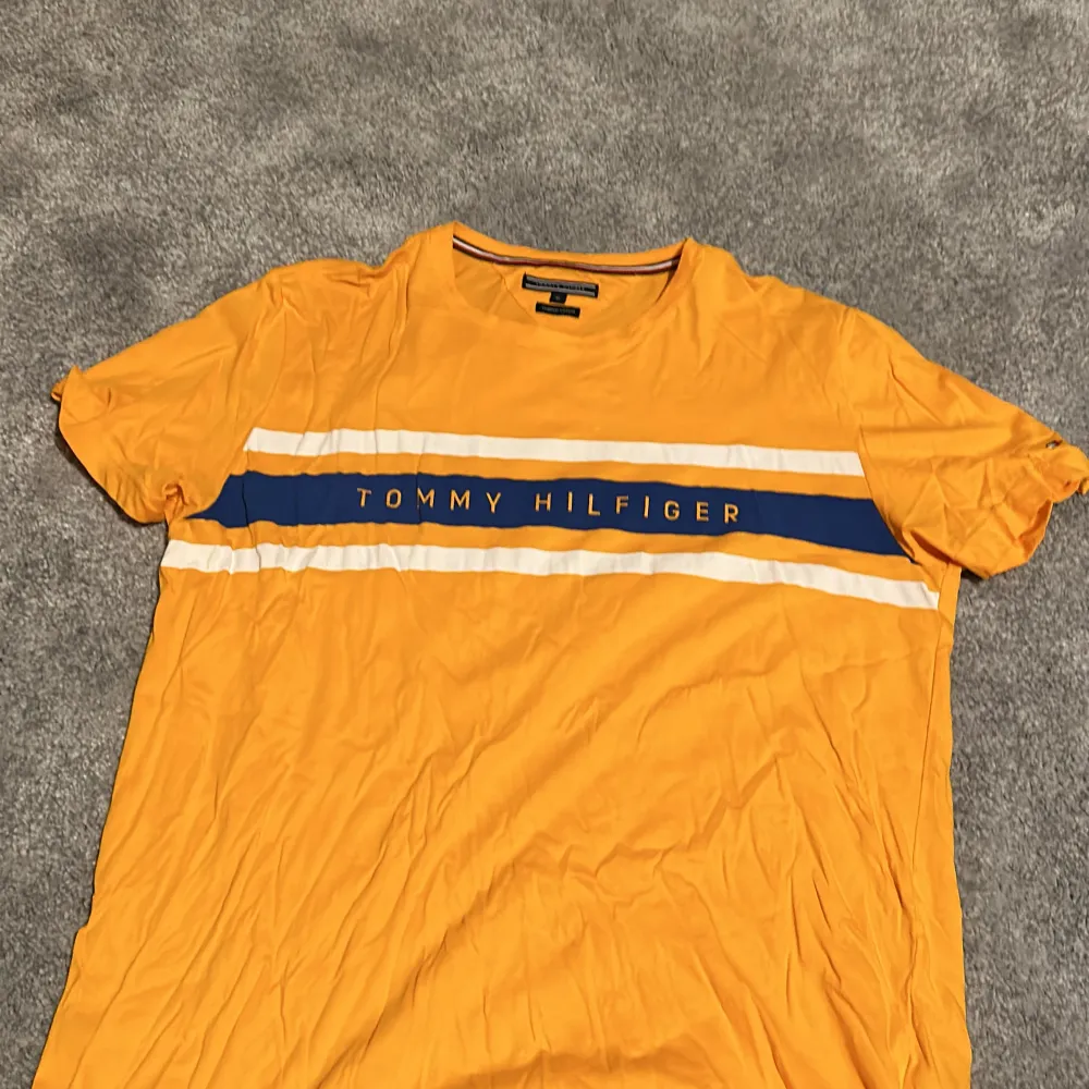 Säljer en snygg orange t-shirt från Tommy Hilfiger i ekologisk bomull. Den har korta ärmar och ett coolt blått och vitt band över bröstet med märkesnamnet. Perfekt för sommaren!. T-shirts.