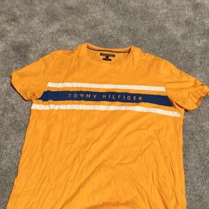Orange t-shirt från Tommy Hilfiger - Säljer en snygg orange t-shirt från Tommy Hilfiger i ekologisk bomull. Den har korta ärmar och ett coolt blått och vitt band över bröstet med märkesnamnet. Perfekt för sommaren!