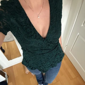Mörkgrön spetsblus - Säljer en superfin mörkgrön spetsblus med v-ringning och korta ärmar. Blusen har ett vackert mönster och en smickrande passform som passar perfekt till både vardag och fest. Perfekt för höst och vår!