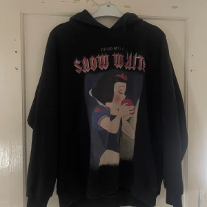 Svart Snow White hoodie från Pull&Bear - Säljer en svart hoodie från Pull&Bear med ett coolt Snow White-tryck på framsidan. Den har en stor och mysig huva och är perfekt för höst och vinter. Materialet är mjukt och bekvämt, vilket gör den idealisk för chill dagar. Passar perfekt för alla Disney-fans!