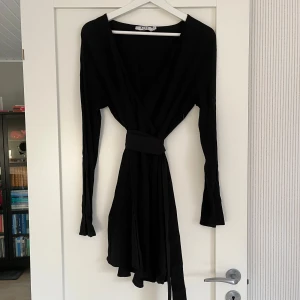 Svart omlottklänning NA-KD - Svart, långärmad, omlottklänning med knyte i midjan från NA-KD i storlek 36. Använd vid ett tillfälle. Klänningen går till halva låret (jag är 170cm).