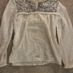 Vit blus med broderad detalj - Säljer en superfin vit blus med långärmad design. Den har en vacker broderad detalj i svart på bröstet och runt halsen. Materialet känns lätt och luftigt. Blusen har en lös passform och är i nyskick. skicka prisförslag vill bli av med den snabbt!💞