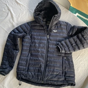 Svart pufferjacka från 8848 Altitude - Säljer en svart pufferjacka från 8848 Altitude. Den är perfekt för kalla dagar med sin vadderade design och har en praktisk huva. Jackan har en smal passform och är sparsamt använd. Superbra för både höst och vinter! 