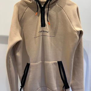 Beige hoodie från c'est normal - Säljer en snygg beige hoodie från c'est normal. Den har en dragkedja vid halsen och svarta detaljer på fickorna. Perfekt för en chill dag eller när du vill vara bekväm men ändå se stilren ut.
