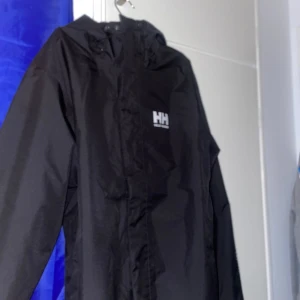 Svart regnjacka från Helly Hansen - Säljer en svart regnjacka från Helly Hansen med Helly Tech-skydd. Jackan har en huva och dragkedja framtill. Perfekt för regniga dagar och håller dig torr och bekväm. 🖤Jackan är aldrig använd, säljer pga att den var för stor på mig. Priset kan diskuteras vid snabb affär