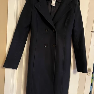 Svart kappa från Patrizia Pepe - Elegant svart kappa från Patrizia Pepe med dubbelknäppning och klassisk krage. Perfekt för kyligare dagar med sin stilrena design och långa ärmar. Tillverkad i Italien, vilket garanterar hög kvalitet och stil.