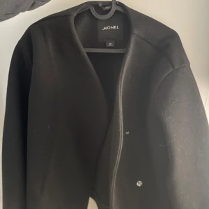 Svart kappa från Monki - Snygg svart kappa från Monki med en stilren design. Jackan har en enkel knäppning framtill och en klassisk krage. Perfekt för att hålla stilen i kallare väder.