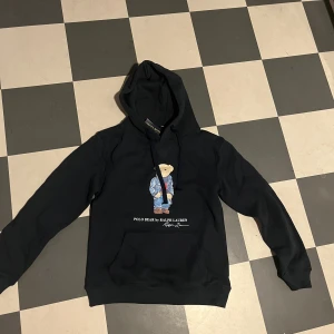 Svart hoodie från Ralph Lauren med björnmotiv - Säljer en svart hoodie från Ralph Lauren med ett charmigt björnmotiv på framsidan. Tröjan har långa ärmar och en klassisk huva med dragsko. Perfekt för en avslappnad stil.