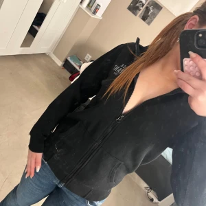 Svart hoodie från S.B. Grant - Snygg svart hoodie från S.B. Grant med dragkedja och fickor framtill. Perfekt för en avslappnad stil. Den har en normal passform och långa ärmar.