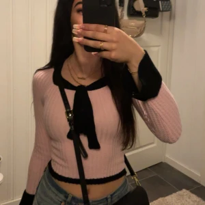 Rosa ribbad tröja med svarta detaljer - Säljer en söt rosa ribbad tröja med svarta detaljer. Tröjan har långa ärmar och en knytning framtill. Den är croppad och har en öppen rygg. Perfekt för en trendig look! Aldrig använt då den inte är min stil💕