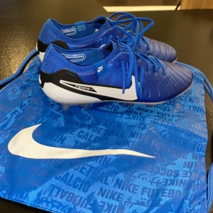 Blå fotbollsskor från nike tiempo elit - Snygga blå fotbollsskor från Nike med vit sula och svarta detaljer. Skorna har snörning och kommer med en matchande blå påse med Nike-logga. Perfekta för fotbollsplanen!