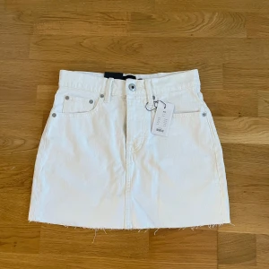 Vit kort jeanskjol från Lager 157 - Snygg kort vit jeanskjol  från Lager 157 med rå kant och klassisk jeansstil. OANVÄND.  Kjolen har fem fickor och en knapp och dragkedja framtill. Perfekt för en stilren look. Köpt för 150kr