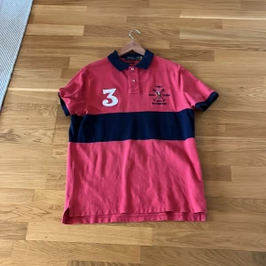 Röd och marinblå pikétröja från Ralph lauren  - Snygg röd pikétröja med marinblått band och stora vita siffror på fram- och baksidan. Tröjan har en klassisk krage och korta ärmar med mörka kanter. Perfekt för en sportig look!