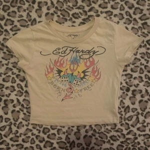  Ed Hardy topp med tryck - Säljer en beige topp från Ed Hardy med ett färgglatt tryck av flammor, tärningar och texte på framsidan. Baksidan har en enkel Ed Hardy-logga. T-shirten har korta ärmar och en rund halsringning.