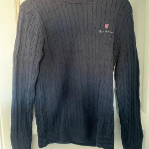 Mörkblå kabelstickad tröja från Bondelid - Snygg mörkblå kabelstickad tröja från Bondelid med broderad logga på bröstet. Tröjan har långa ärmar och ribbade muddar vid ärmslut och nederkant. Perfekt för kyligare dagar! 