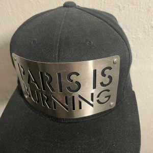 En Paris is burning caps den är metal  - Snygg svart keps från Karl Alley med en unik metallplatta framtill med texten 'PARIS IS BURNING'. Skärmen är grön på undersidan och kepsen är tillverkad av 85% akryl och 15% ull.