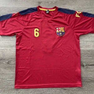 Röd fotbollströja med nummer 6 - Säljer en röd fotbollströja med korta ärmar och nummer 6 tryckt i gult. Tröjan har FCB-logga på bröstet och mörkblå detaljer på axlarna. Perfekt för fotbollsfans!
