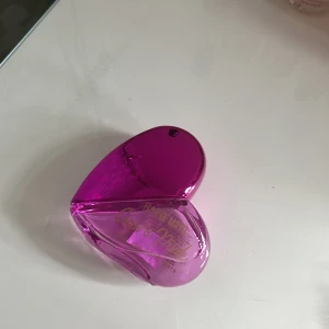 Parfym i hjärtformad flaska - En charmig parfym i en hjärtformad flaska med en glansig rosa färg. Flaskan har en unik design som gör den till en fin detalj på sminkbordet. Perfekt för den som älskar en söt och lekfull stil.