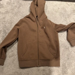 Brun hoodie från Ralph Lauren - Säljer en snygg brun hoodie från Ralph Lauren med dragkedja och huva. Tröjan har långa ärmar och två praktiska fickor framtill. Perfekt för en avslappnad stil.