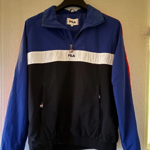 Fila jacka  - Säljer en snygg flerfärgad vindjacka från FILA med dragkedja och hög krage. Jackan har en blå överdel, svart nederdel och en vit rand över bröstet med FILA-loggan. Ärmarna är långärmade med en röd detalj längs sidan. Perfekt för en sportig look!