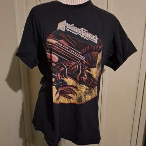 Svart Judas Priest t-shirt från Gildan - Cool svart t-shirt från Gildan med ett stort tryck av Judas Priest och texten 'Screaming for Vengeance'. Perfekt för fans av bandet! T-shirten är kortärmad och gjord i bomull för en bekväm passform.