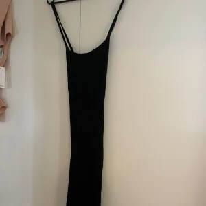 Svart långklänning - Elegant svart långklänning med smala axelband. Perfekt för en stilren look. Klänningen har en figurnära passform som framhäver silhuetten. Skims liknande i modellen. Korsad i ryggen. 