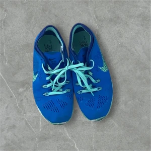 Blå Nike sneakers - Säljer ett par blå Nike sneakers med ljusgröna detaljer och snörning. Skorna har ett ventilerande mönster och en flexibel sula, perfekt för träning eller vardagsbruk.