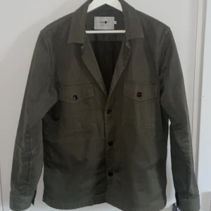 Grön overshirt från NN07 - Snygg grön overshirt från NN07 med knappar framtill och två bröstfickor. Perfekt för lager-på-lager-stil. Den har en klassisk krage och långa ärmar, vilket gör den idealisk för både vår och höst. Nyskick från märket nn07 storlek S kan gå ner i pris vid snabb affär skriv vid minsta fråga eller fundering!