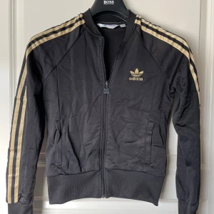 Svart Adidas jacka/tröja med guldiga ränder - Säljer en svart Adidas jacka med ikoniska guldiga ränder längs ärmarna och Adidas-logga på bröstet. Jackan har dragkedja framtill och ribbade muddar vid ärmslut och nederkant. Perfekt för en sportig look!