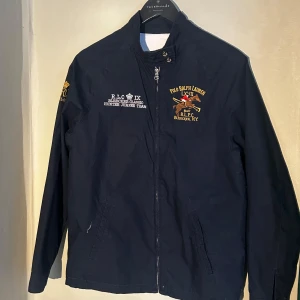 Mörkblå jacka - Polo Ralph Lauren - Polo Ralph Lauren jacka med broderade detaljer på bröstet och ärmen. Saknar tagg i nacken.