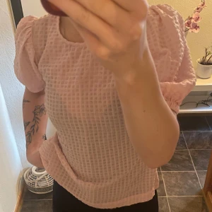 Rosa blus med puffärmar - Säljer en söt rosa blus med puffärmar och ett rutigt mönster. Perfekt för en stilren look. Blusen har en rund halsringning och är tillverkad i ett lätt och luftigt material.