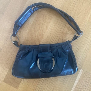 Svart axelväska från Venezia - Svart axelväska från Venezia i skinn. Väskan har en justerbar axelrem och en dekorativ metallspänne framtill. Perfekt för att bära dina nödvändigheter med stil. Genuine leather made in Italy. 