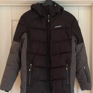 Svart och grå skidjacka från Icepeak i stl 164 - Säljer en svart och grå skidjacka från Icepeak i strl 164 (13-14 år) med avtagbar huva, snölås och liftkortsficka på ena ärmen. Jackan har två praktiska fickor med dragkedjor framtill. Väldigt sparsamt använd och i gott skick.