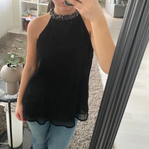 Svart ärmlös topp med pärldetaljer - Snygg svart ärmlös topp med hög hals och vackra pärldetaljer runt kragen. Toppen har en elegant plisserad design och är perfekt för en stilfull look. Passar bra till både jeans och kjol.