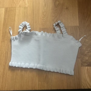 Vit ribbad tubtopp från Zara - Säljer en söt vit ribbad tubtopp från Zara med volangdetaljer. Perfekt för varma sommardagar. Toppen har elastiska axelband som ger en bekväm passform.
