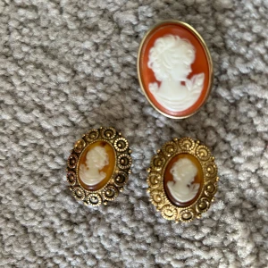Vintage cameosmycken - Säljer ett set med vintage cameosmycken bestående av en brosch och ett par örhängen. Broschen har en vit profil på en orange bakgrund, medan örhängena har en liknande design med en guldfärgad ram. Perfekt för att ge en klassisk touch till din stil.