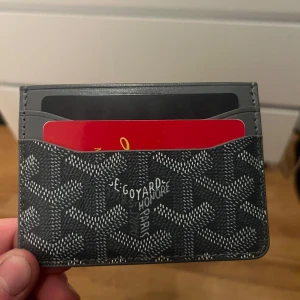 Grå korthållare från Goyard - Säljer ett stilrent grått kortfodral från Goyard med deras ikoniska mönster. Perfekt för att hålla dina kort organiserade och lättillgängliga. Kommer i en elegant grön förpackning med Goyards logotyp i silver.