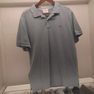 Ljusblå pikétröja från GANT - Säljer en stilren ljusblå pikétröja från GANT i regular fit. Tröjan har en klassisk krage och korta ärmar, med GANT-loggan broderad på bröstet. Perfekt för en avslappnad och snygg look.