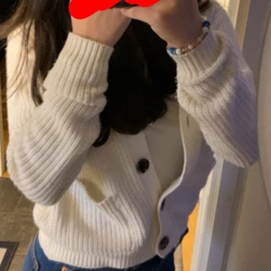 Vit stickad kofta från H&M - Säljer en mysig vit stickad kofta från H&M med stora knappar framtill. Koftan har långa ärmar och två fickor fram, perfekt för kyliga dagar. Passar både till jeans och kjol för en avslappnad look.