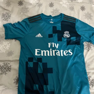Blå Real Madrid fotbollströja - Säljer en blå Real Madrid fotbollströja från Adidas med korta ärmar. Tröjan har Fly Emirates som sponsorlogga och ett coolt mönster i svart. På baksidan finns namnet Ronaldo och nummer 7 tryckt i vitt. Pris kan diskuteras!