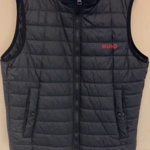 Svart dunväst från Hugo boss - Snygg svart dunväst från Hugo boss (st M) med röd logga på bröstet. Västen har en dragkedja framtill och två fickor med knappar. Perfekt för lager-på-lager under kyligare dagar. Hör av dig vid frågor