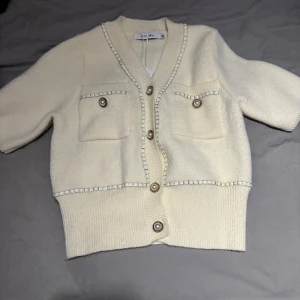 Beige kofta från Knit Mix - Säljer en stilren beige kofta från Knit Mix med korta ärmar och dekorativa knappar framtill. Koftan har två fickor på bröstet och ribbade kanter. Perfekt för en elegant och avslappnad look.